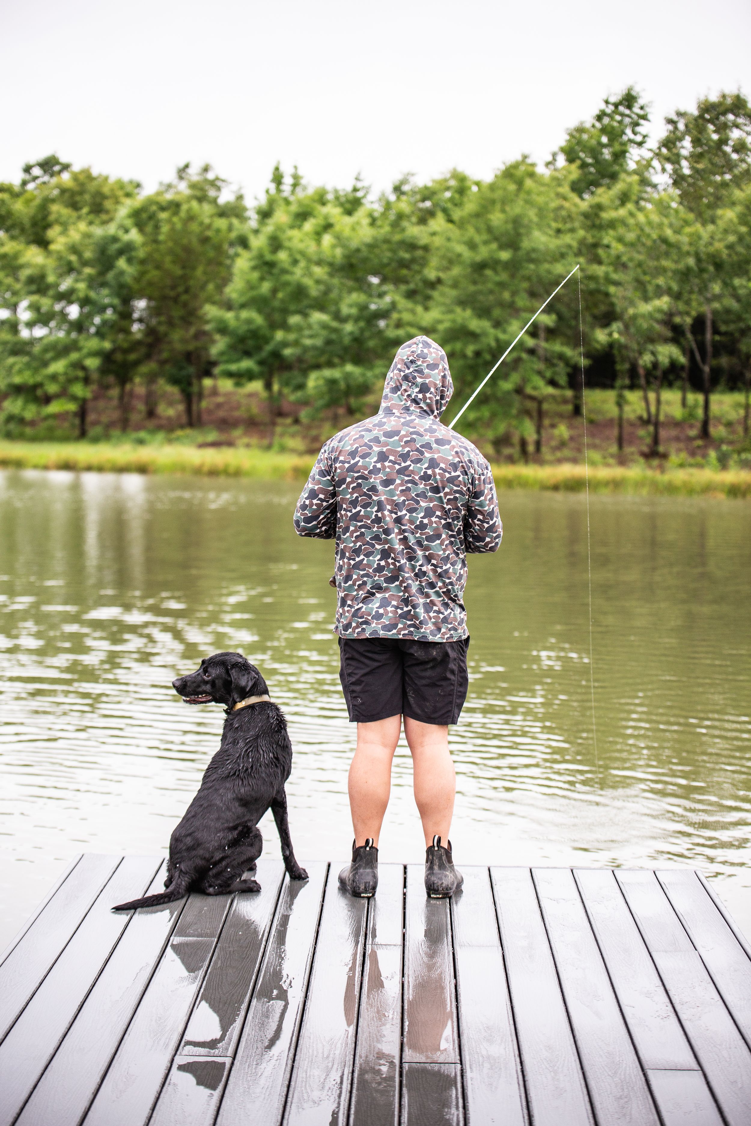Person in floral shirt fishing from wooden dock with dog on peaceful lake.