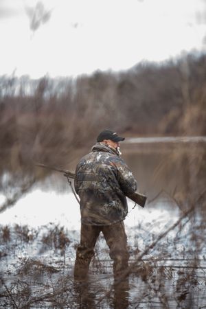 Hunter in camouflage gear wading through flooded marshland with a shotgun.
