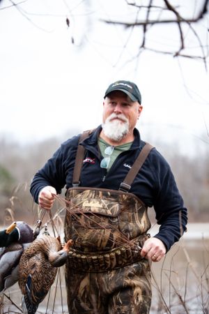 Duck hunter in camouflage waders displays harvested waterfowl in natural setting.