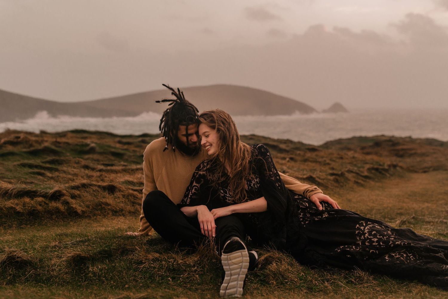 A couple sits close together on a grassy hilltop at sunset with mountains and misty coastline in the background.