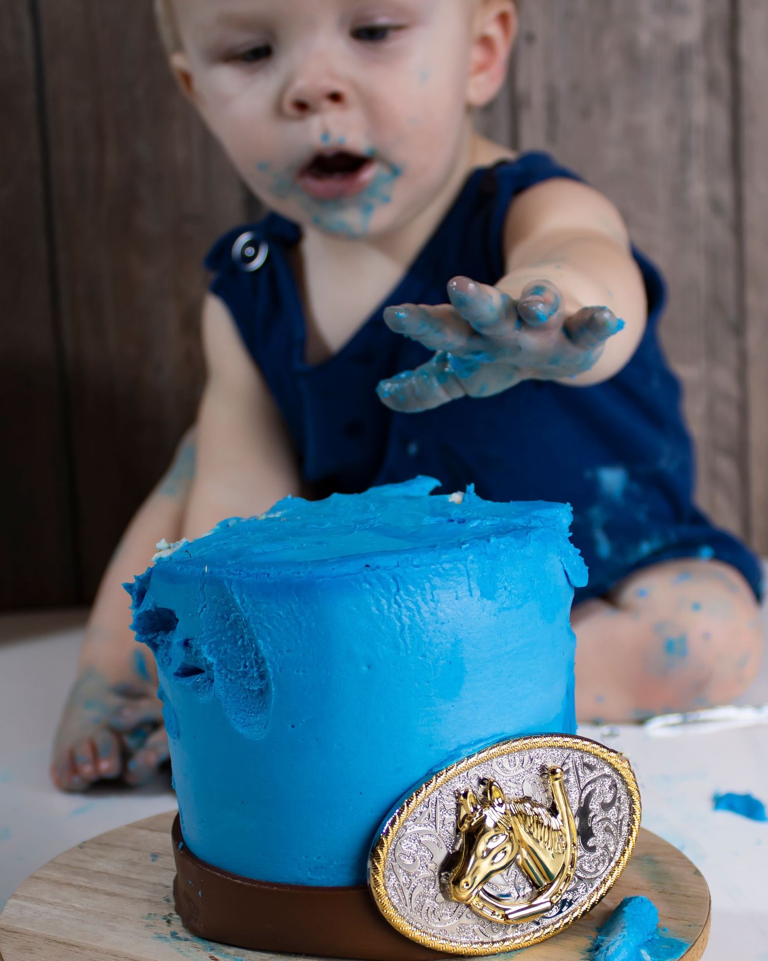 A baby boy reaches for a bright blue cake. The boy and cake are covered in messy frosting
