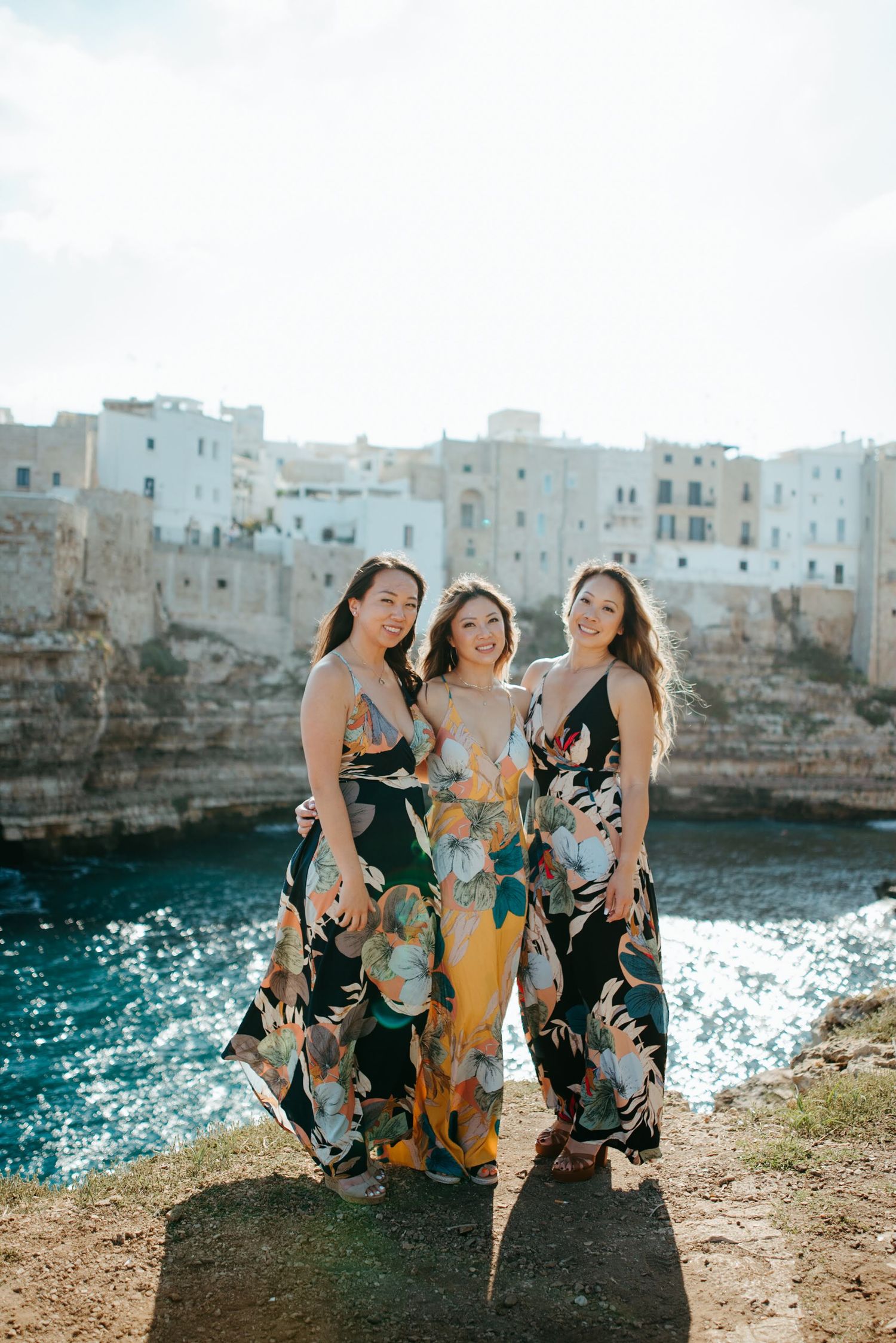 Three women in floral dresses sit together by the sparkling Mediterranean Sea.