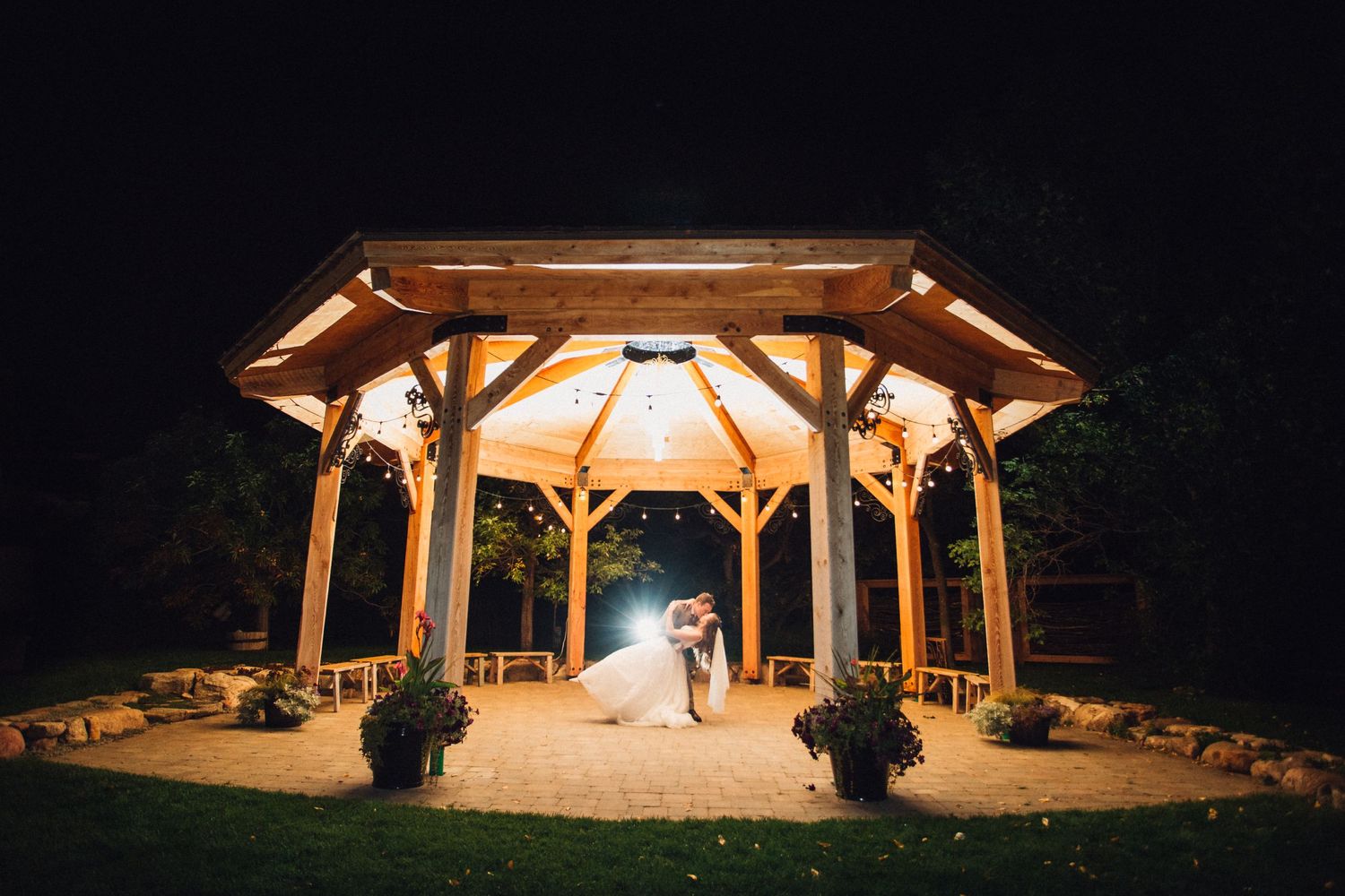 Wooden gazebo illuminated at night with elegant figures in wedding attire.