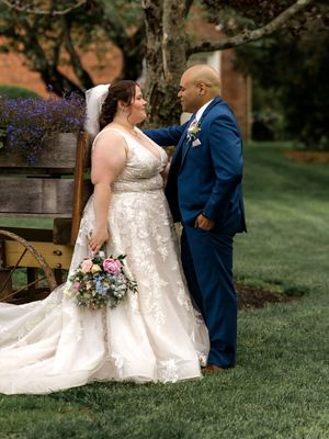 A romantic wedding couple stands on a lawn with purple flowers and trees in the background as the bride holds a floral bouquet.