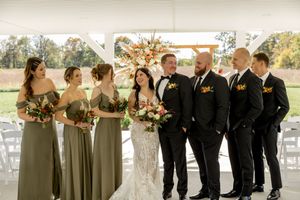 Wedding party in sage green dresses and black tuxedos pose together under a wooden arch at an outdoor venue.