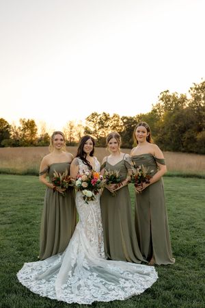 Wedding party poses together in olive green dresses and white lace gown against golden sunset in grassy field.