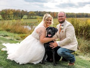 A couple in wedding attire poses with their black Labrador retriever dog in a scenic outdoor autumn setting.