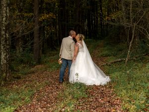 A couple in wedding attire shares romantic moments in a wooded area with fallen autumn leaves on the ground.