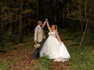 A couple in wedding attire dances and celebrates together in a scenic wooded outdoor setting during autumn.