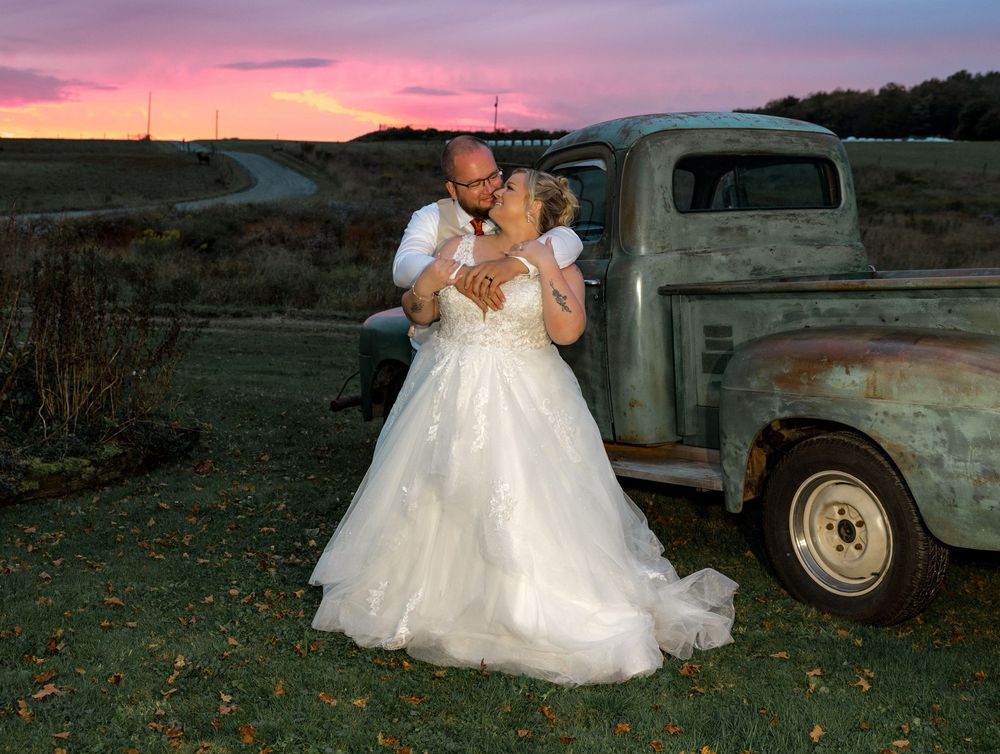 A bride in a white wedding dress poses at sunset with a vintage truck in a rustic outdoor setting.