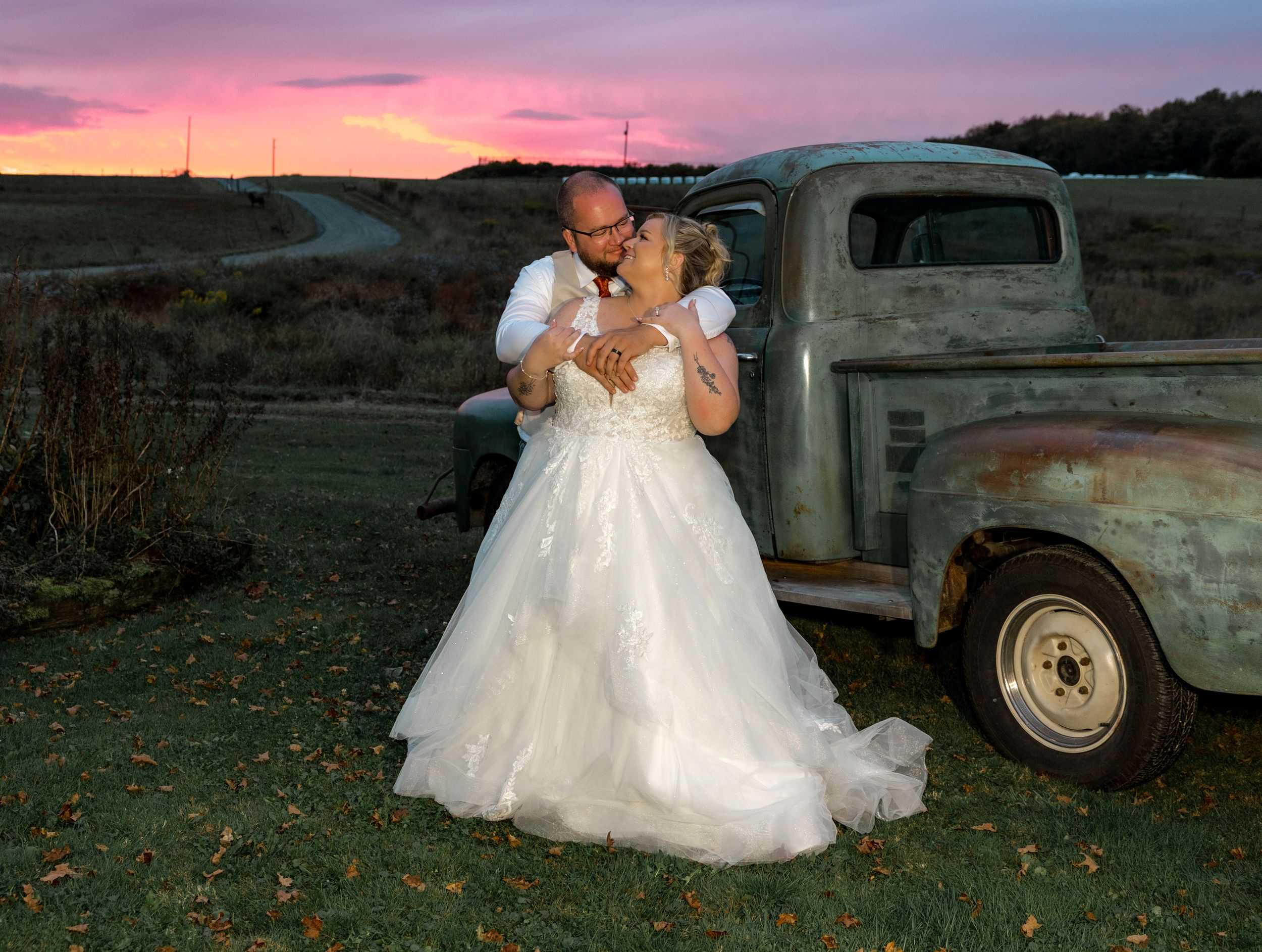A bride in a white wedding dress poses at sunset with a vintage truck in a rustic outdoor setting.