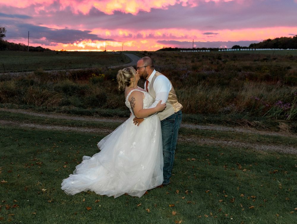 A couple in wedding attire shares a romantic moment at sunset in a grassy field with a pink and purple sky.