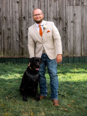 A person in a beige suit and jeans poses with a black dog in front of a wooden fence in a backyard setting.