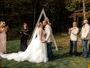 A bride and groom share a kiss during an outdoor fall wedding ceremony under a wooden arch.