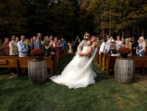 Wedding guests gather around rustic wooden barrels at an outdoor ceremony venue during sunset.