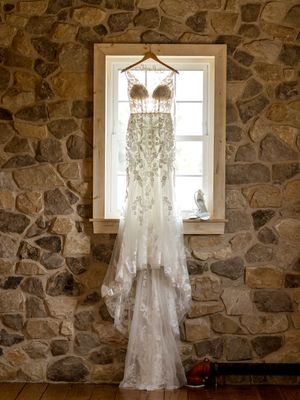 A delicate lace wedding dress hanging in a window against a rustic stone wall creates an ethereal bridal scene.