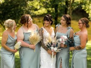 Bridesmaids in soft sage green dresses gather around bride in white lace gown holding bouquets in sunny garden setting.