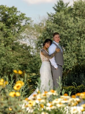 A wedding couple stands together in a wildflower garden filled with yellow daisies and lush greenery on a sunny day.