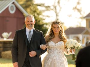 A bride and father walk arm-in-arm down a sunlit path during a wedding ceremony at golden hour.