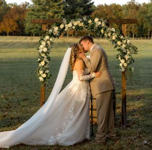 Romantic outdoor wedding ceremony setup with white floral arch and wooden benches in autumn field at sunset.