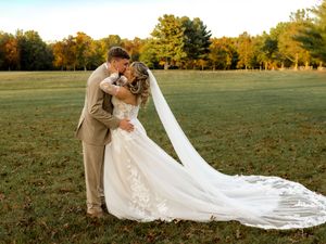 Romantic outdoor wedding scene with couple embracing as bride's long veil trails behind on grass.