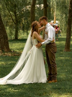 Couple sharing an intimate kiss under tall trees in a wooded outdoor wedding setting with flowing white dress and veil.