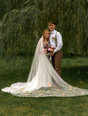 Wedding couple poses in front of weeping willow trees with bride's flowing cathedral veil spread on autumn grass.
