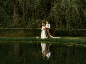 Two people in wedding attire stand together beneath a weeping willow tree reflected in a serene pond at sunset.