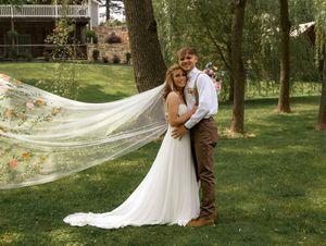 A couple shares a romantic moment outdoors with a flowing long wedding veil catching the breeze in a lush green garden setting.