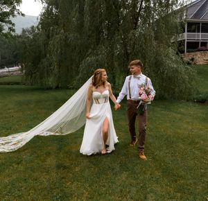 A couple walks through a grassy field during a wedding photoshoot with a flowing white veil in the breeze.