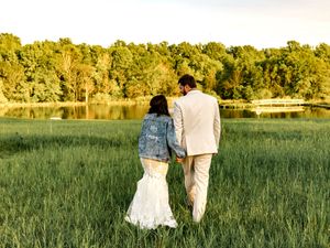 A couple sits in a grassy meadow at golden hour with the sun setting behind tall trees in the distance.