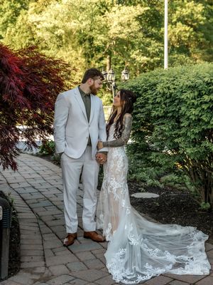 A wedding couple walks together on a stone path in a garden setting wearing a grey suit and lace gown.
