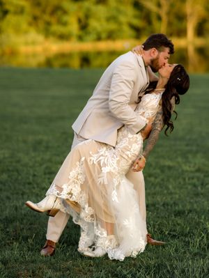 A romantic couple in wedding attire dips and kisses on a green grassy field during sunset in matching white outfits.
