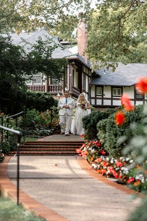 A scenic wedding moment on brick steps leading to a Tudor-style house with red flowers blooming along the pathway.