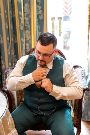 A person in a green vest and white dress shirt adjusts their tie while seated on an elegant patterned chair.