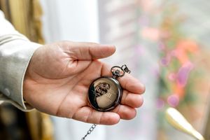 Hand holds an antique pocket watch against a blurred colorful background as light reflects off its metal surface.
