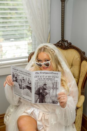 Bride in classic white veil sitting on vintage chair reads different newspaper poses during wedding photoshoot.