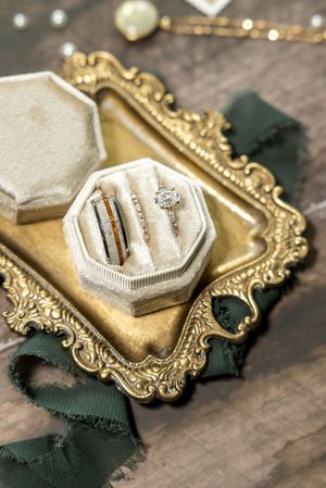 Elegant engagement ring nestled in velvet-lined hexagonal box on ornate gold vintage tray with emerald green ribbon.