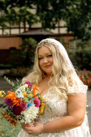 Bride in lace wedding dress holds vibrant autumn-colored bouquet with dahlias and wildflowers in outdoor garden setting.