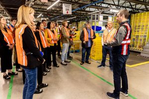 Group of workers in orange safety vests receiving instructions during a warehouse facility tour.
