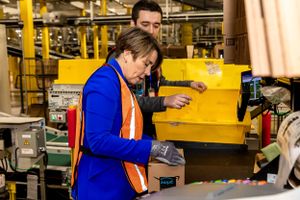 Worker in blue jacket and safety vest operating equipment in a warehouse setting.