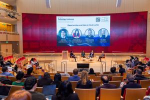 Panel discussion event in modern auditorium with red backdrop and presentation screen.