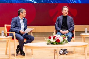 Two presenters having discussion on stage with wooden furniture and red backdrop.