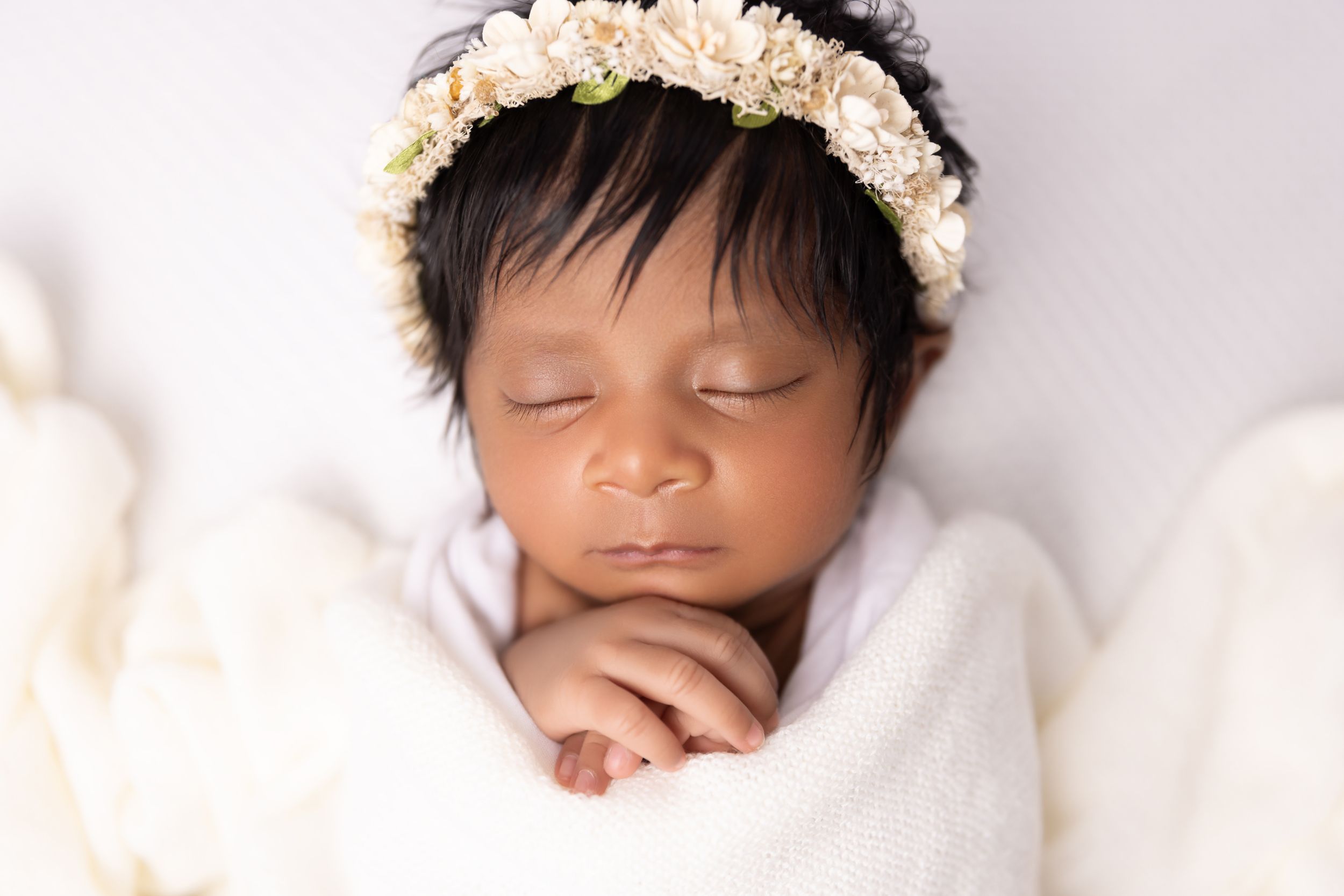 Peaceful sleeping newborn wearing delicate floral headband wrapped in soft white blanket.