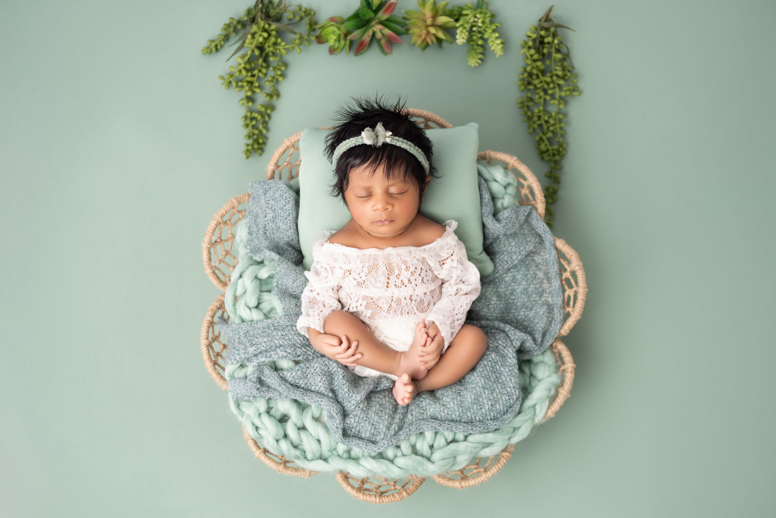 Sleeping infant in white lace dress lying in woven basket with blue blanket and greenery backdrop against mint wall.