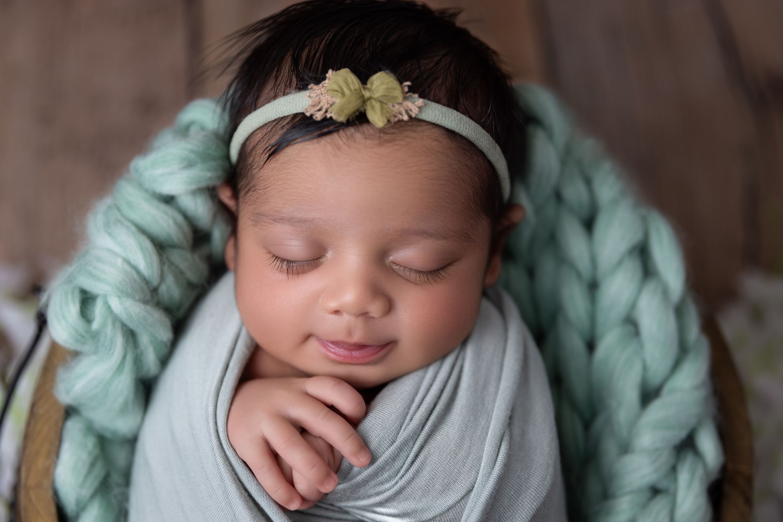 Sleeping newborn swaddled in light gray fabric with floral headband on teal chunky knit blanket background.