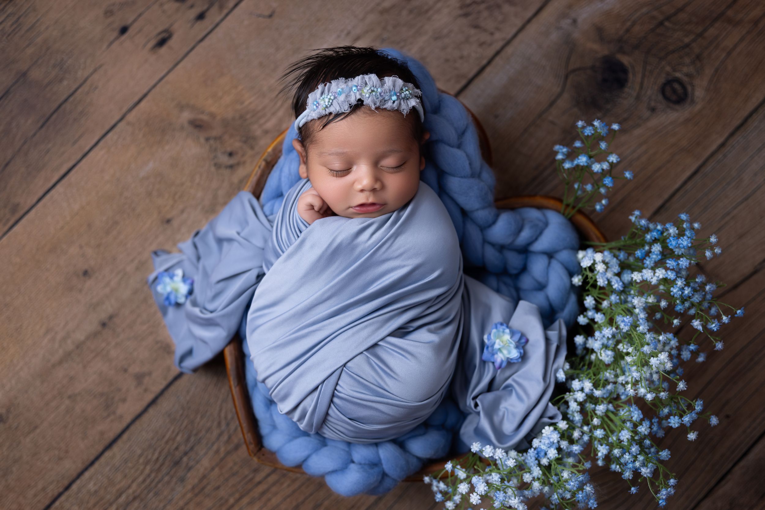 A sleeping baby swaddled in soft blue fabric lies on wooden floors with delicate white flowers scattered nearby.