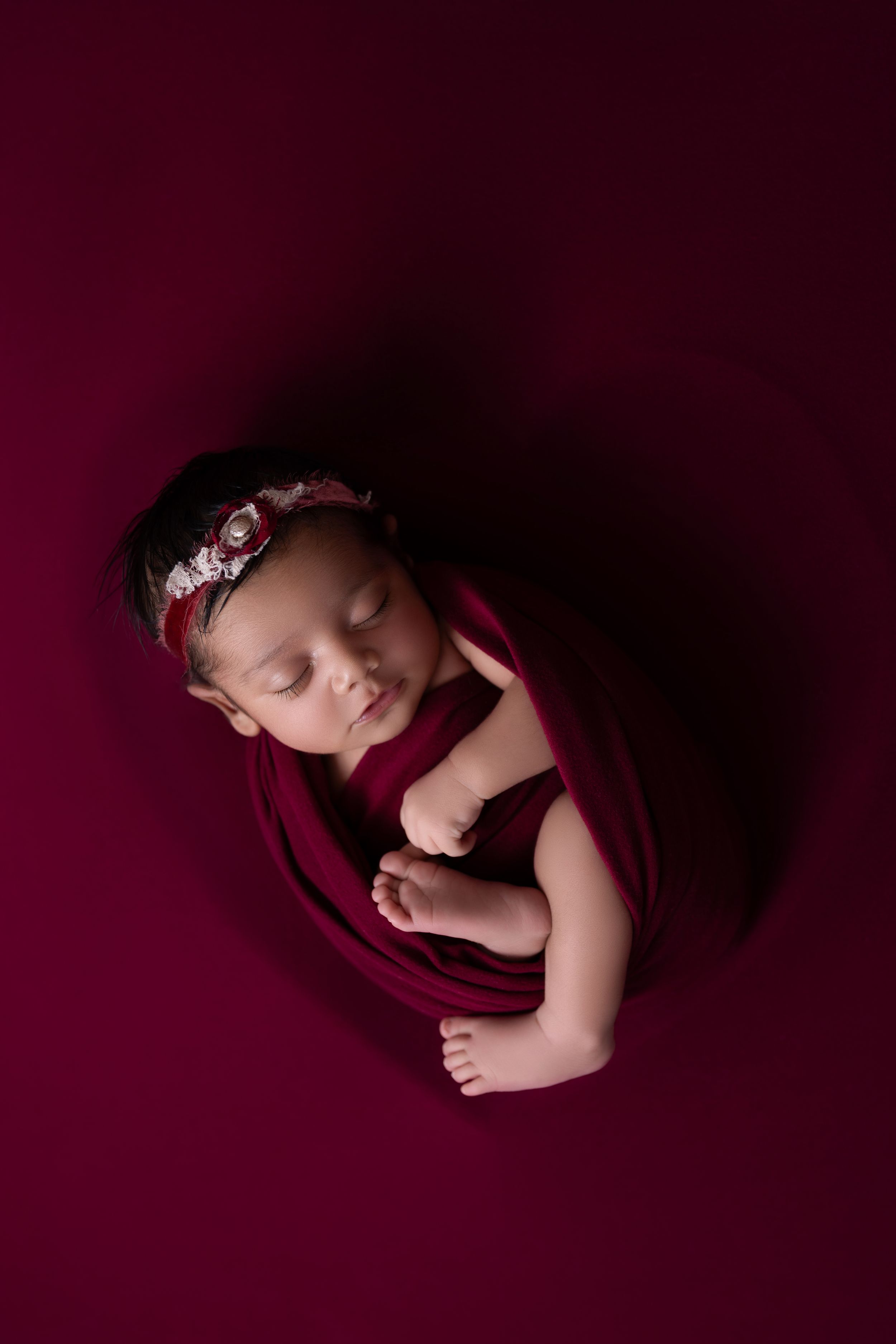 Sleeping newborn baby wearing floral headband curled up on deep burgundy backdrop in artistic portrait.