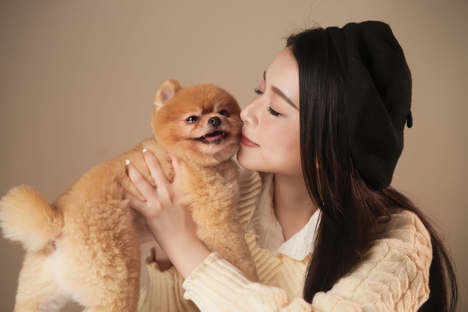 A person with dark hair lovingly cuddles and kisses their fluffy cream-colored Pomeranian dog.