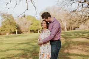 A engaged couple embraces in a sunny park setting with green trees in the background.